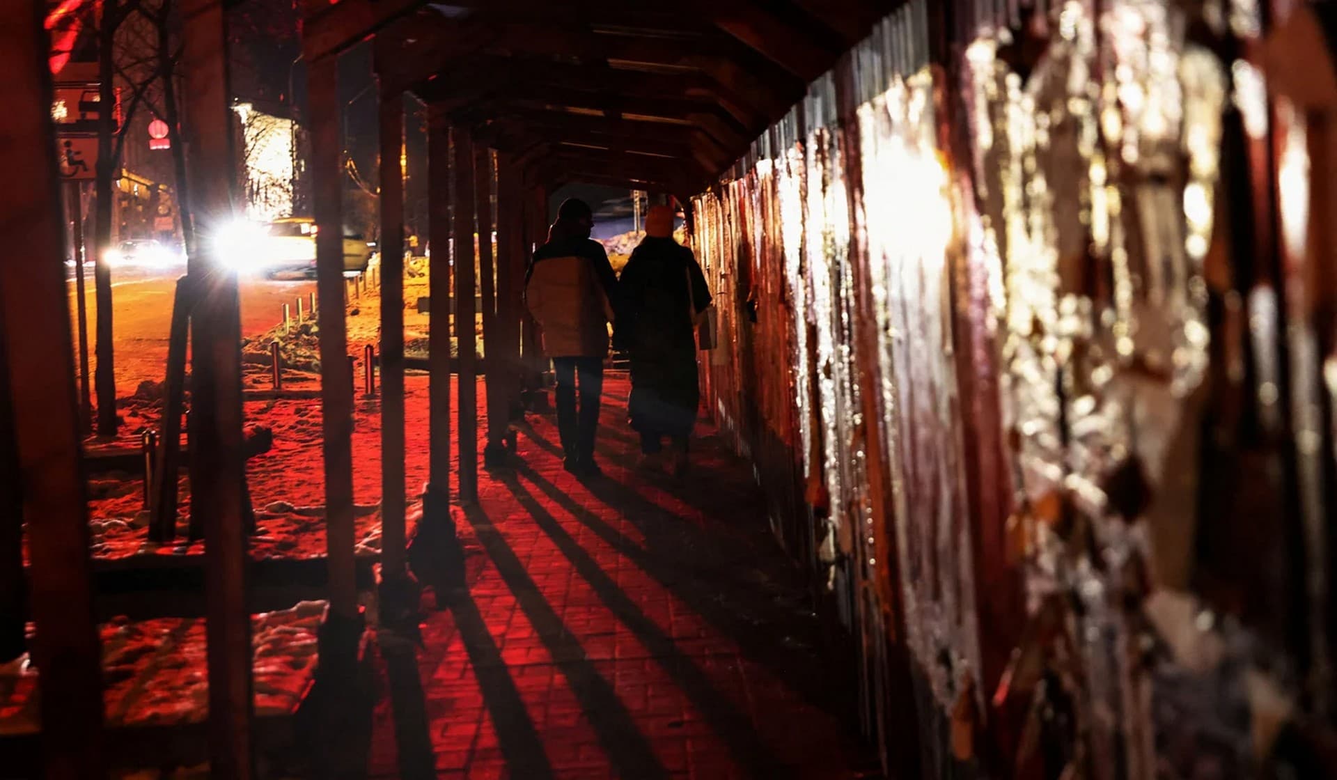 People walk on a sidewalk illuminated by automobile headlights during power outages in Kyiv