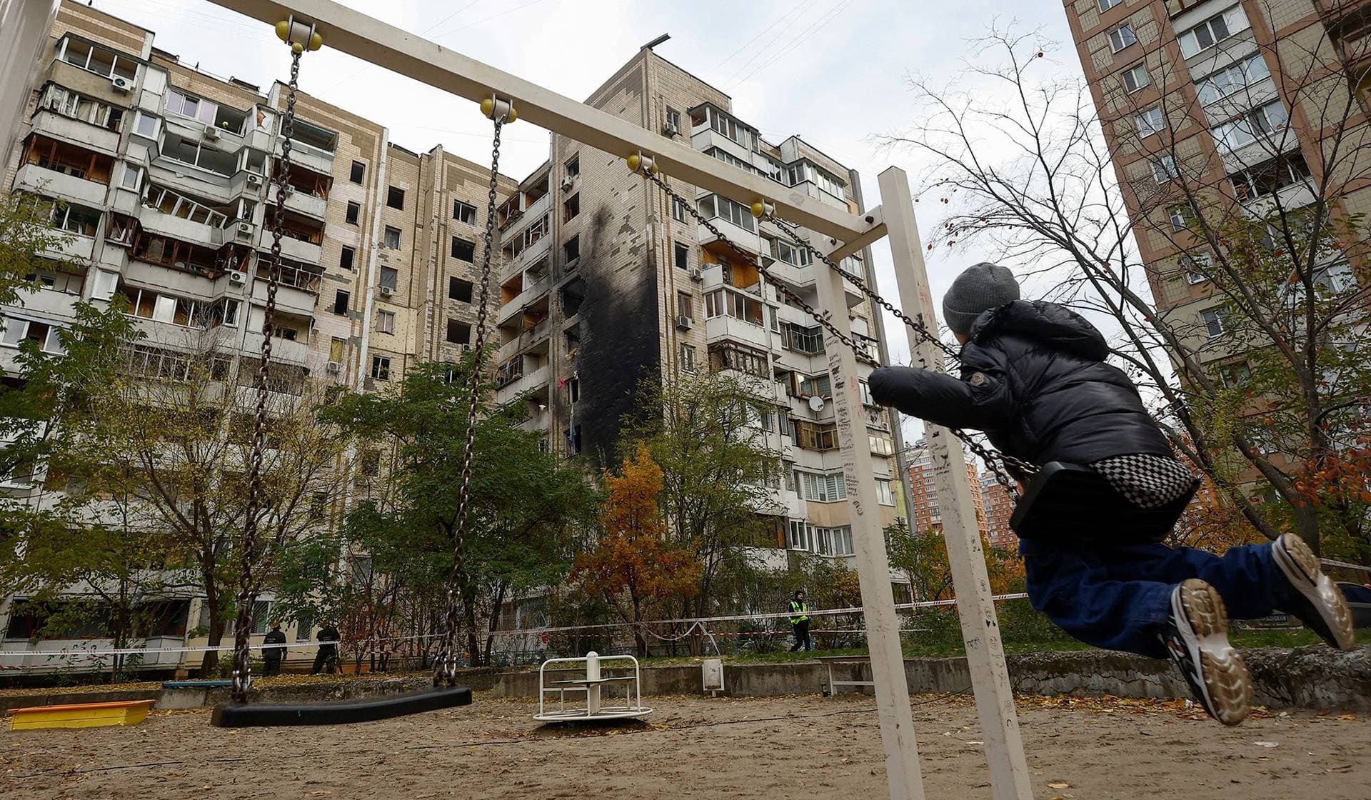 A boy sits on a swing at a playground in front of an apartment building damaged during an overnight Russian drone strike in Kyiv